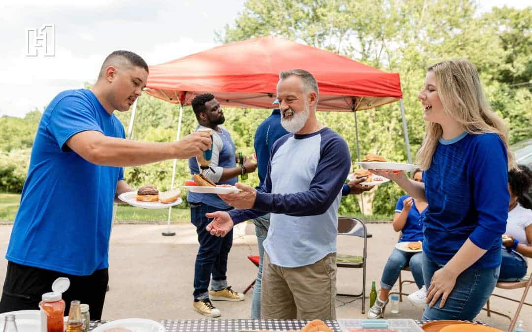 Three adults at tailgate in parking lot for sporting event being served hot dogs and burgers